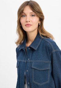 Denim shirt in dark blue with a collared neckline, button front, two chest pockets, and contrasting stitching details. Smooth texture.