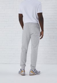 Pantalones de chándal grises con un ajuste entallado, cinturilla elástica y tobillos ajustados, combinados con zapatillas deportivas grises y doradas frente a un fondo de ladrillo blanco.