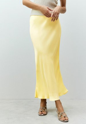 Femme portant une longue jupe jaune en satin, un haut beige en maille sans manches, une bague en or et des sandales beige à brides, debout sur un sol gris.