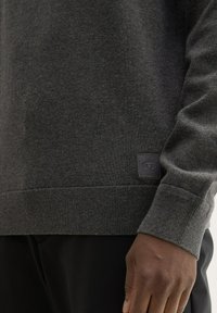 Ciemnoszary sweter o miękkiej fakturze, z prążkowanymi mankietami i dołem, z małą naszywką logo na dole po lewej stronie.