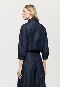 Navy blue blouse with three-quarter sleeves, featuring a high collar, textured fabric, and gold button accents on the cuffs. Back view displayed.