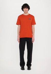 Red cotton t-shirt with a crew neck and short sleeves, featuring a black rectangular logo patch on the upper left. Paired with black pants.