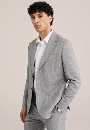 Noor mees seisab kandes helerohelist triibulist ülikonda valge särkega, käsi taskus, neutraalne taust.