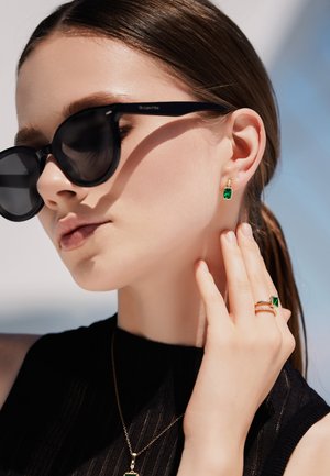 Goudkleurige sieraden met smaragdgroene stenen. Het model draagt stud oorbellen, een ring en een hanger aan een ketting, aangevuld met zwarte zonnenbril.