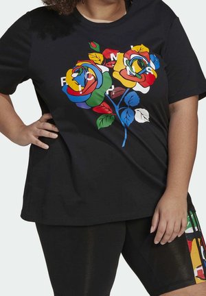 Persona con curvas que lleva una camiseta negra con rosas abstractas y coloridas y texto blanco parcial, combinada con pantalones cortos negros con un panel lateral multicolor.