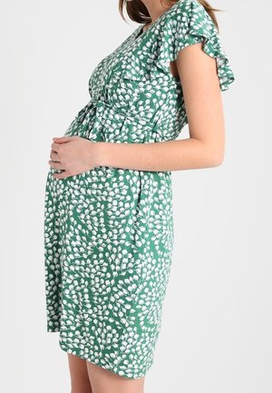 Kobieta w ciąży w zielonej sukience w biały kwiatowy wzór delikatnie trzymająca się za brzuch, stojąca na tle jednolitego jasnego tła.