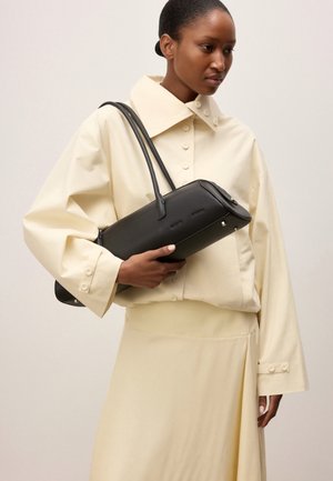 Femme en blouse crème et jupe tenant un sac à main en cuir noir avec deux poignées d'épaule sur un fond uni.