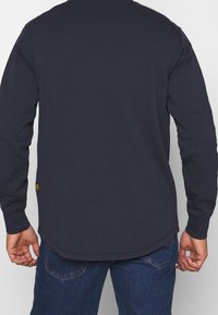 Marinblå långärmad tröja med rund hals och rundad nederkant. Tillverkad av bomull med en etikett på den undre sidan. Texturerat tyg, avslappnad passform.