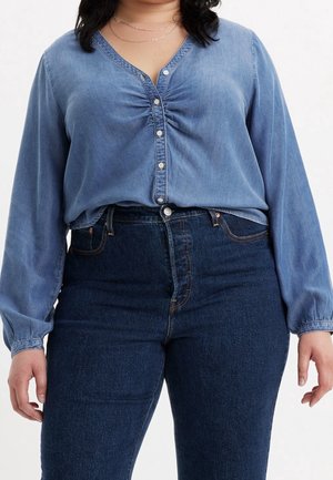 Kvinde iført en blå denim knapbluse med opsamlet brystdetalje og højtaljede mørkeblå jeans, stående foran en hvid baggrund.