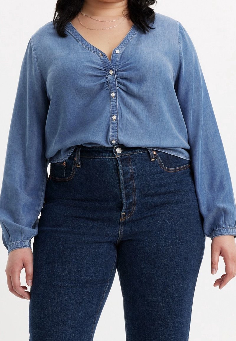 Femme portant un chemisier en denim bleu boutonné avec un détail froncé au niveau de la poitrine et un jean bleu foncé taille haute, debout devant un fond blanc.