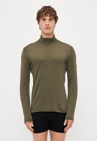 Olivovo zelená dlhá rukávová mikina na zips vyrobená z ľahkého materiálu, so stojacím golierom a úzkym strihom, nosená nad čiernymi športovými šortkami.