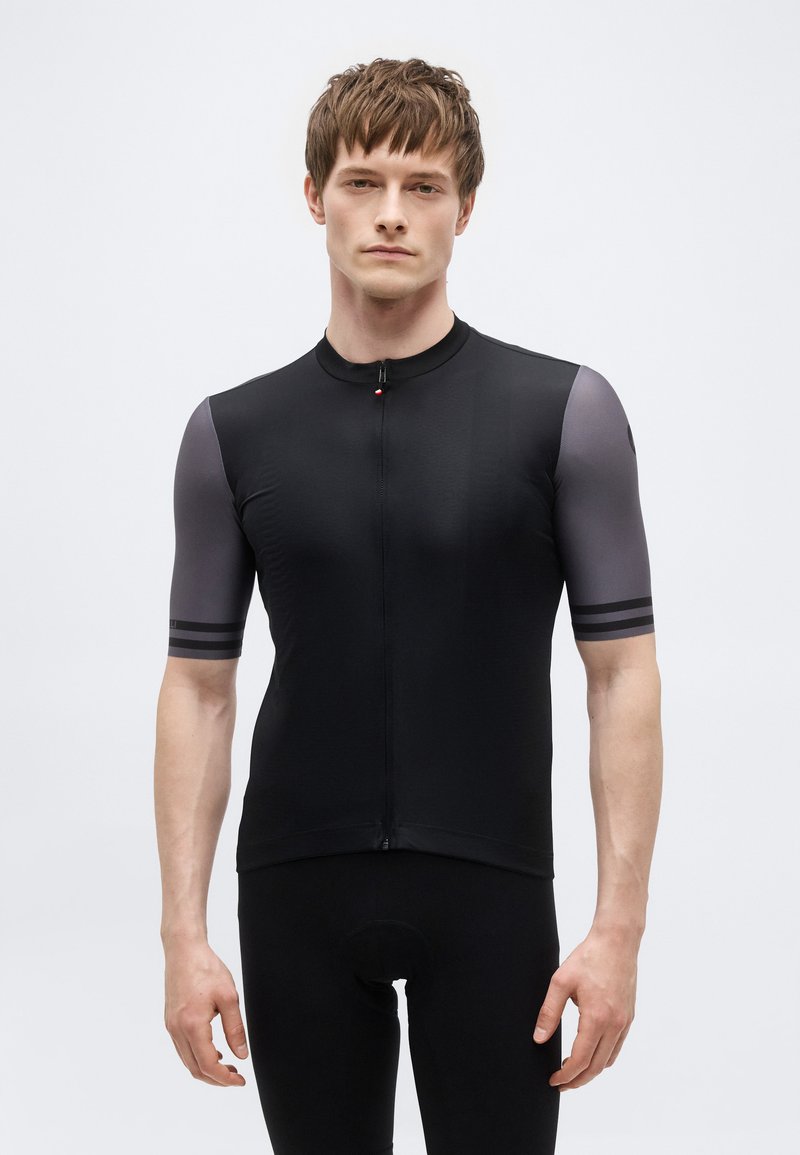 Giovane uomo con una maglia da ciclismo nera a maniche corte con maniche grigie e una cerniera frontale, in piedi davanti a uno sfondo chiaro e uniforme.