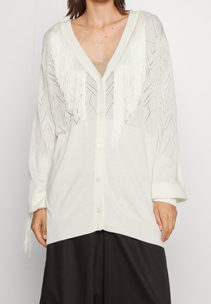 Cardigan blanc en maille avec détails à franges sur la poitrine et les manches, porté sur un haut beige et un pantalon noir.