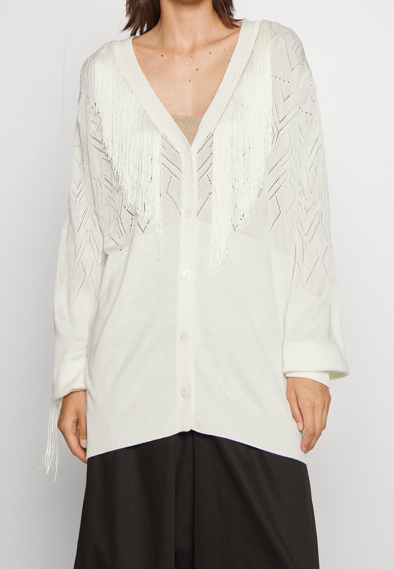 Cardigan blanc en maille avec détails à franges sur la poitrine et les manches, porté sur un haut beige et un pantalon noir.