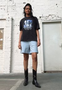 Svart grafisk t-shirt med konstverk, ljusblå texturerade shorts och svarta western-stil stövlar. Enkel bakgrund med en dörr.