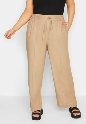 Pantalon beige à jambes larges avec taille à cordon et poches latérales, porté avec des sandales noires et un top noir sans manches, montré sur un modèle partiel.