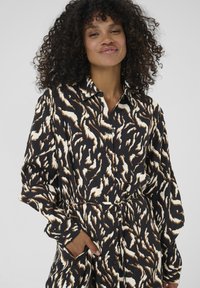 Abito shirt nero con un motivo astratto in crema e marrone, maniche lunghe, chiusura a bottoni frontale e una cintura coordinata. Tessuto dalla consistenza liscia.