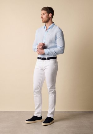 Homme portant une chemise bleu clair boutonnée, pantalon blanc, ceinture noire et chaussures noires à enfiler, debout devant un fond beige.