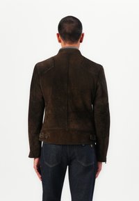 Jachetă din suede maro cu un design ajustat, având detalii matlasate la umeri și catarame laterale, privită din spate pe un fundal alb.