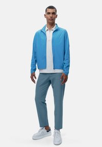 Világoskék cipzáras dzseki fehér pólóing felett, világoskék nadrággal és fehér sportcipővel párosítva. Egyszerű, modern dizájn lélegző anyagból.