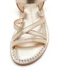 Sandal de metal dorado con diseño de tiras cruzadas, textura suave, detalles cosidos en la suela y punta abierta, que refleja la luz.