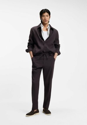 Young man with black hair wearing dark brown relaxed suit, white undershirt, and dark brown slip-on shoes, standing with hands in pockets.