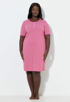 Nainen seisoo paljain jaloin, yllään vaaleanpunainen polvipituinen mekko, jossa on kuvioita, lyhyet hihat ja napit kaula-aukossa.