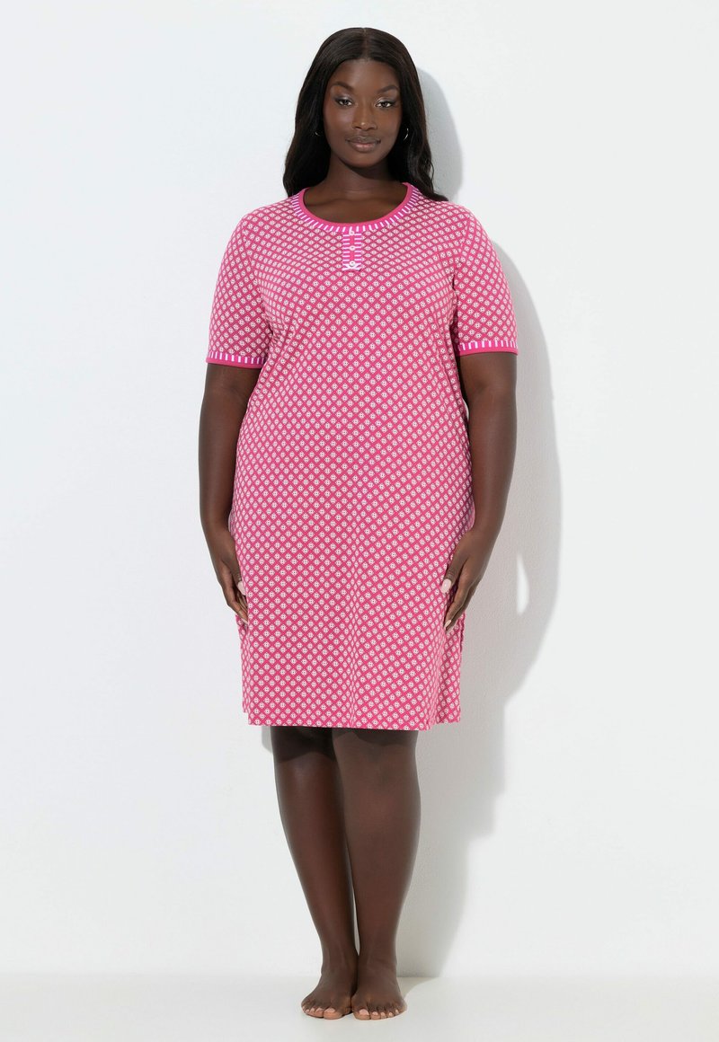 Mujer de pie descalza con un vestido rosa con estampado, de longitud hasta la rodilla, manga corta y detalles de botones en el escote.