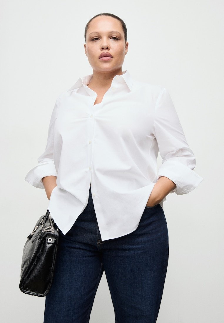 Sebevědomá žena v bílé košili na knoflíky a tmavých džínách, ruce v kapsách, drží černou koženou kabelku na levé straně.