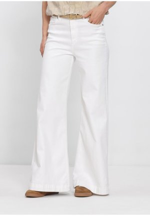 Person trägt hoch geschnittene, weit geschnittene weiße Hose mit beigem Gürtel und braunen Schuhen, steht vor einem schlichten weißen Hintergrund.