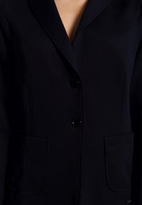 Blazer bleu marine en tissu lisse, avec un col à revers, deux poches avant et trois boutons noirs pour la fermeture.
