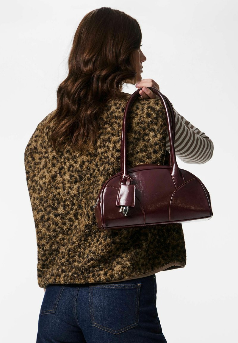 Burgundia nahast käekott ümarate vormidega, lühikeste sangade ja väikese sildiga. Kotil on sile tekstuur ja kõverad detailid.