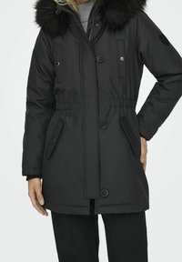 Parka noire matelassée avec une capuche en fausse fourrure, taille ajustable, fermeture boutonnée croisée et poches latérales. Texture lisse.