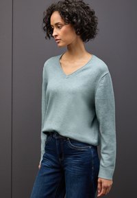 Hellblauer, strukturierter Strickpullover mit V-Ausschnitt und langen Ärmeln, kombiniert mit dunkelblauen Jeans. Der Hintergrund ist ein neutrales Grau.