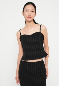 Svart avskåret tank topp med tynne stropper, med en bølgete utringning og vertikale hvite striper. Glatt tekstur, tettsittende silhuett.