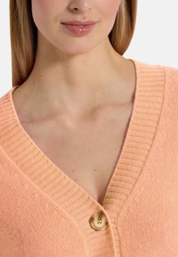 Perzikkleurige gebreide cardigan met een V-halsontwerp en geribbelde afwerking, voorzien van een houten knoopsluiting op de borst, die de zachte textuur benadrukt.