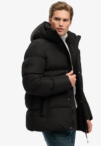 Man in zwart gewatteerd hoodie-jack over een witte trui en jeans, kijkt opzij en past de rits van zijn jack aan met zijn rechterhand.
