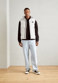 Doudoune légère grise avec col montant, portée sur un polo noir, associée à un jean bleu clair et des baskets noires sur un sol en bois.