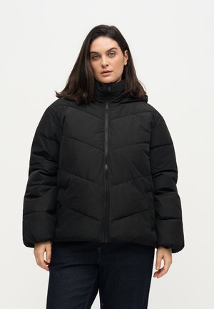 Mujer con cabello oscuro hasta los hombros, vistiendo una chaqueta acolchada negra y vaqueros oscuros, de pie frente a un fondo blanco liso.