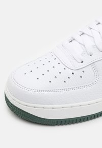 Bílé kožené tenisky s perforovanou špičkou, plochým zavíráním na šňůrky a texturovanou zelenou gumovou podešví. Obsahují silné švy a kulatý design špičky.