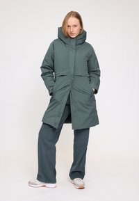 Grüne, isolierte Parka mit hohem Kragen, Druckknopfverschlüssen und zwei vorderen Taschen. Über weiten Cord-Hosen und Sportschuhen getragen.