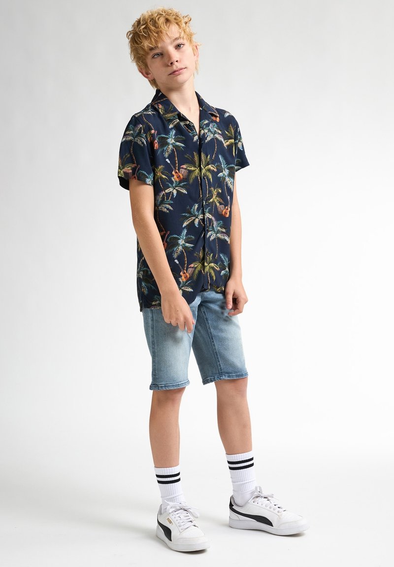 Cămașă bleu închis cu mânecă scurtă, cu imprimeu de palmieri, purtată cu pantaloni scurți din denim de un albastru deschis. Asortată cu adidași albi și șosete cu dungi. Stil casual.