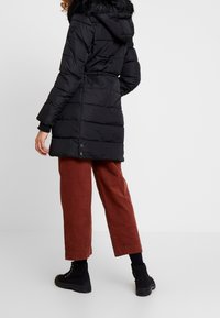 Chaqueta negra acolchada con capucha, con borde de piel sintética, diseño acolchado y cintura ajustable con cordón. Se lleva sobre pantalones anchos de color óxido.