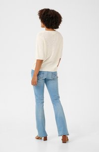 Haut beige clair à manches courtes associé à un jean évasé bleu. Les jeans présentent un motif de couture subtil. Le mannequin porte des sandales à talons carrés marron.
