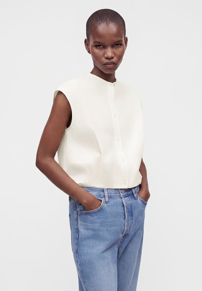 Femme aux cheveux courts portant une chemise blanche sans manches boutonnée et un jean bleu, les mains dans les poches, sur un fond uni.