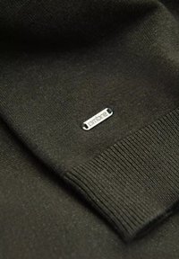 Maglione verde scuro con polsini a coste e un'etichetta metallica argentata con il testo "Ombre", che indica il dettaglio del marchio. Texture liscia, design classico.