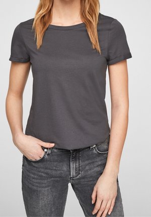 Femme portant un t-shirt gris foncé à manches courtes et un jean noir délavé, une main dans la poche, cadrée du cou jusqu'au milieu de la cuisse.