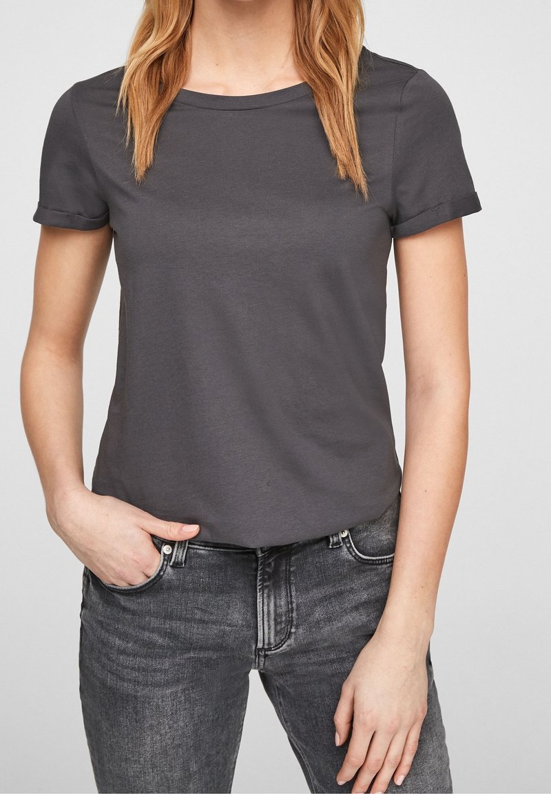 Femme portant un t-shirt gris foncé à manches courtes et un jean noir délavé, une main dans la poche, cadrée du cou jusqu'au milieu de la cuisse.
