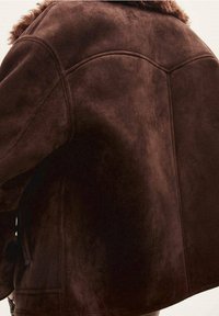 Brown suede jacket featuring a textured finish, curved seams, and a cozy faux fur collar. Accents include top-stitching details.