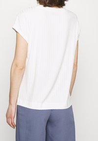 Persona che indossa un top bianco a maniche corte a coste e pantaloni blu larghi, mostrata di spalle su uno sfondo neutro.